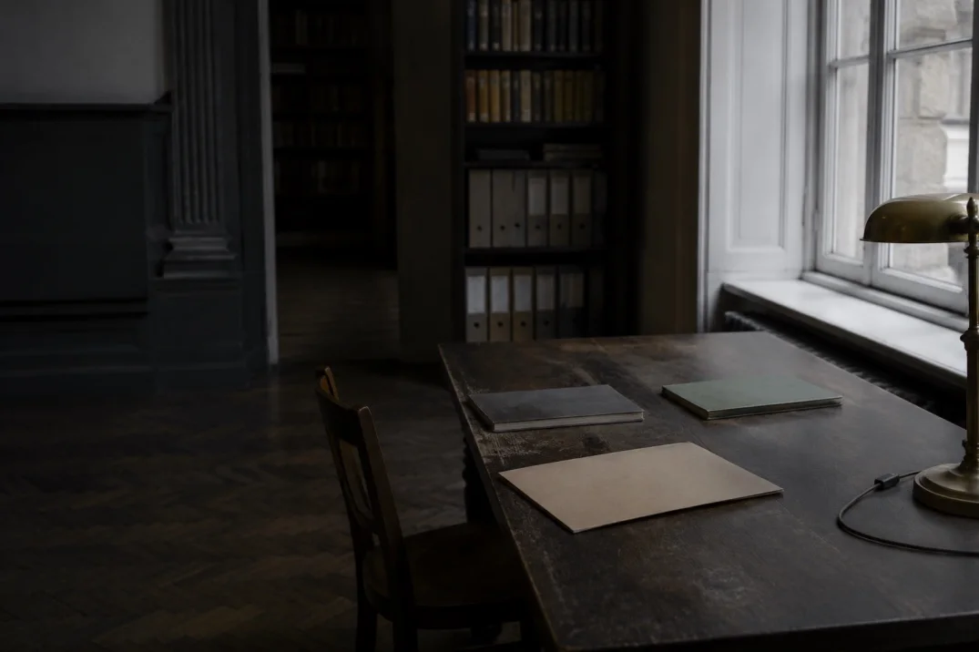 A dark wood desk with two folders and a leather document holder beneath a brass banker's lamp, a library visible through an open doorway.
